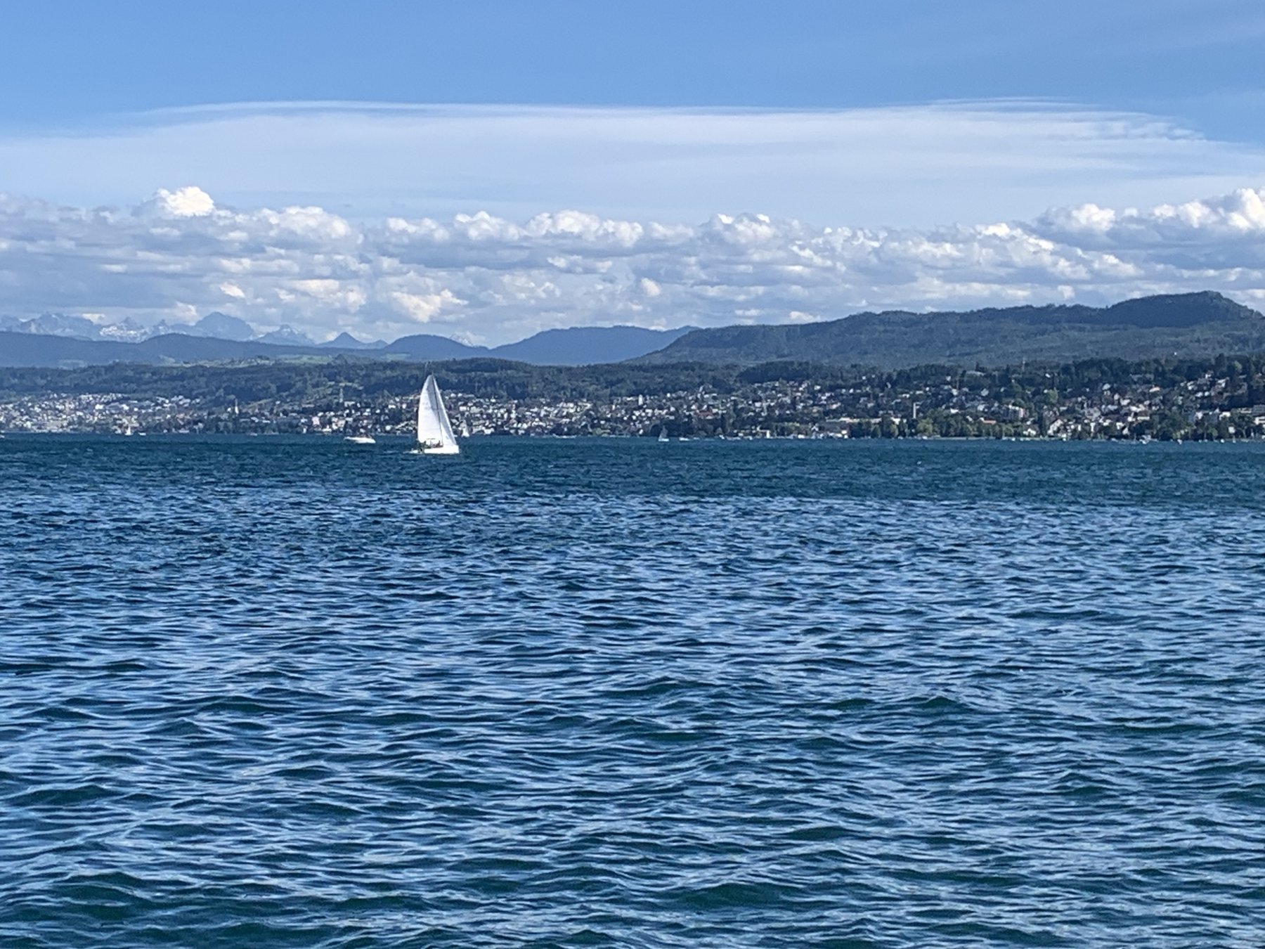 A calm lake scene with a sailboat, hills, and mountains in the distance